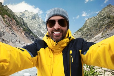 Image of Smiling man in hat and sunglasses taking selfie in mountains