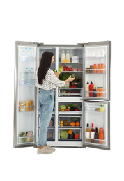 Young woman with bag of groceries near open refrigerator on white background