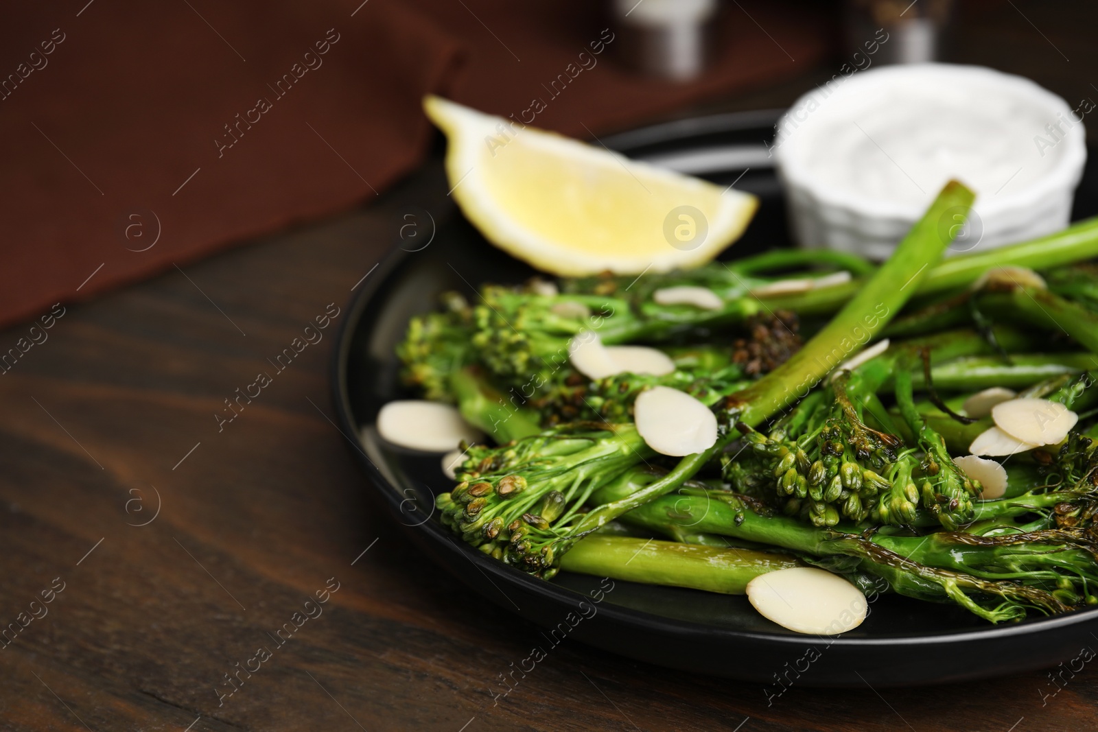 Photo of Tasty cooked broccolini with almonds, lemon and sauce on wooden table, closeup. Space for text