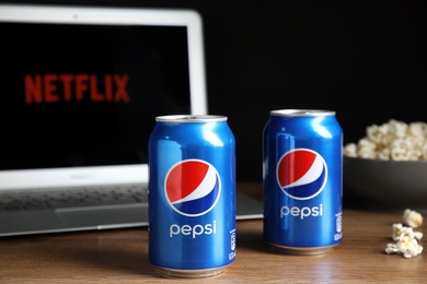 MYKOLAIV, UKRAINE - FEBRUARY 17, 2021: Cans of Pepsi and pop corn near laptop displaying Netflix on wooden table