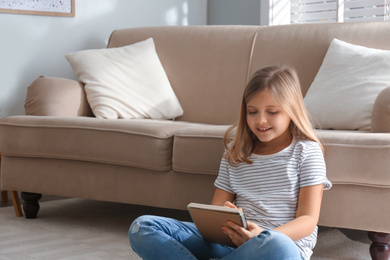 Photo of Cute little girl with notebook near couch at home. Space for text