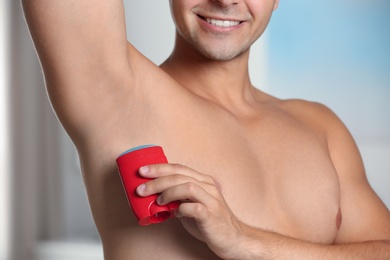 Young man applying deodorant to armpit indoors, closeup