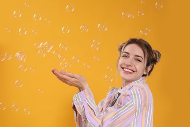 Young woman playing with soap bubbles on yellow background, space for text