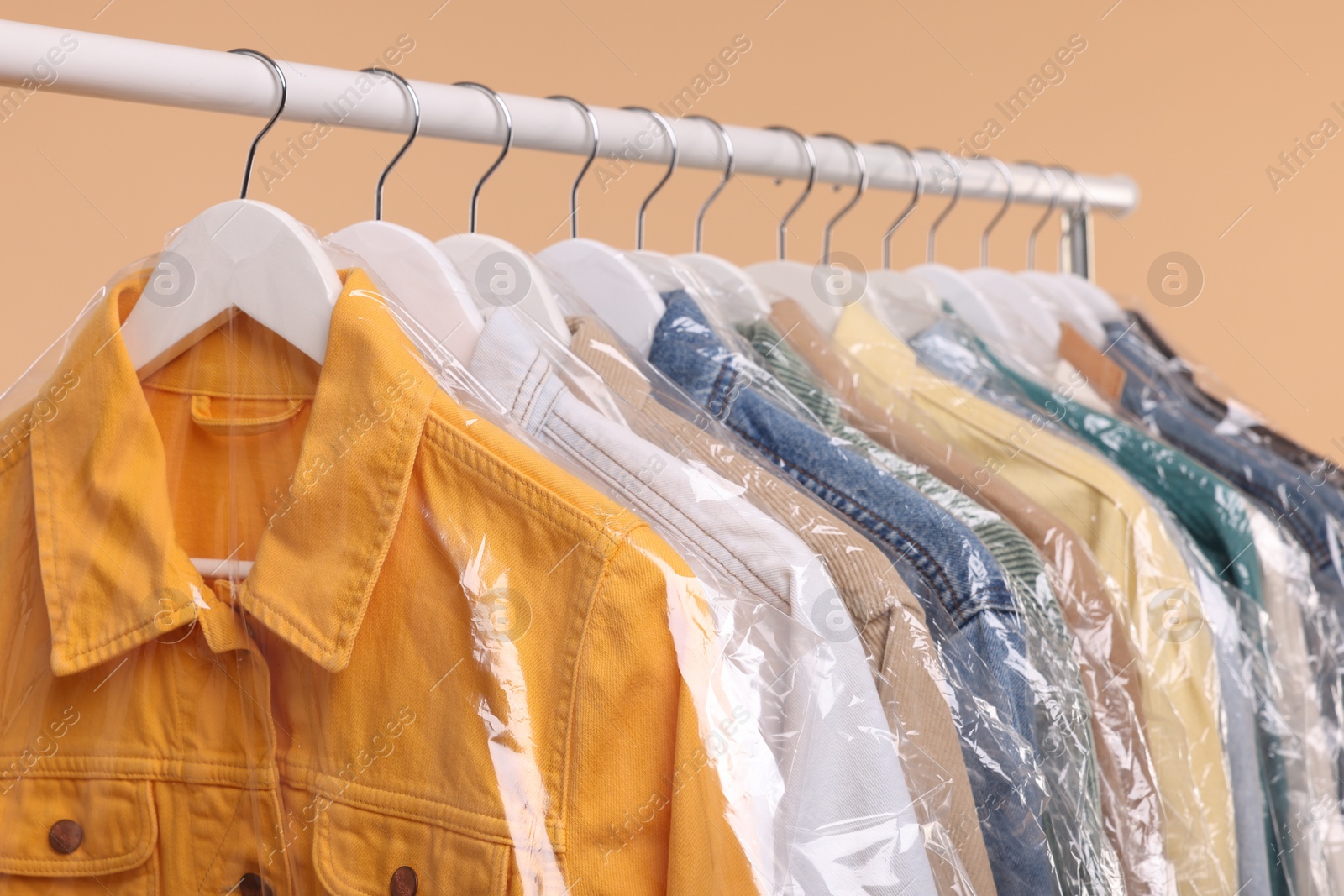 Photo of Dry-cleaning service. Many different clothes in plastic bags hanging on rack against beige background