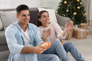 Photo of Happy couple with tangerine near sofa in room decorated for Christmas