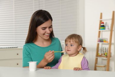 Mother feeding her cute little child with yogurt at white table in room