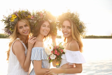 Young women wearing wreaths made of beautiful flowers near river on sunny day
