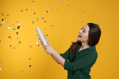 Young woman blowing up party popper on yellow background