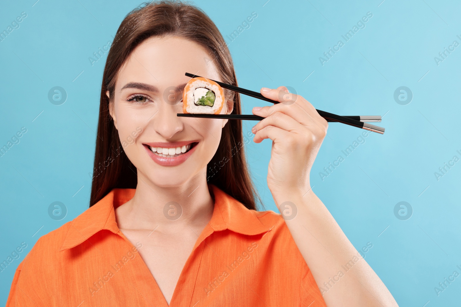 Photo of Funny young woman holding sushi roll with chopsticks on light blue background