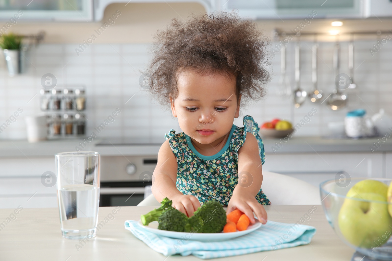 Photo of Cute African-American girl eating vegetables at table in kitchen