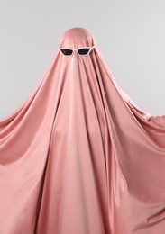 Glamorous ghost. Woman in pink sheet with sunglasses on light grey background