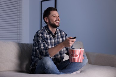 Happy man with popcorn bucket changing TV channels with remote control at home in evening