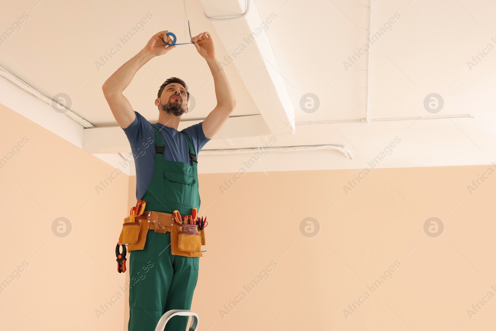 Photo of Electrician in uniform with insulating tape repairing ceiling wiring indoors. Space for text