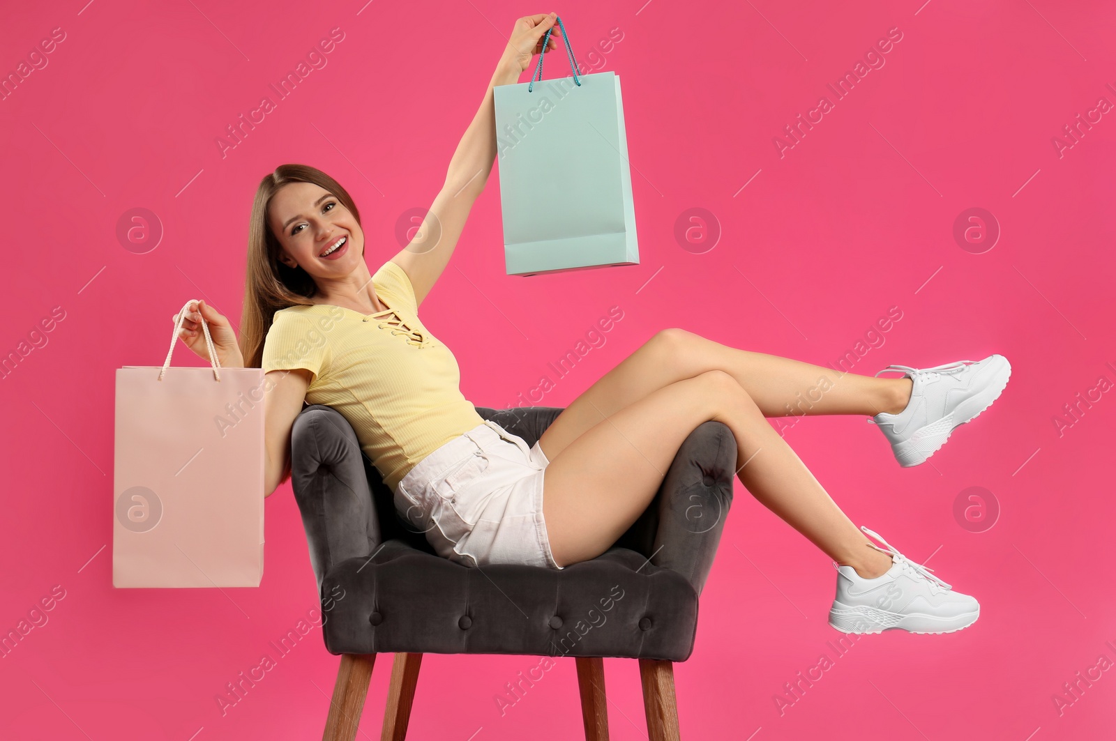 Photo of Beautiful young woman with paper shopping bags in armchair on pink background