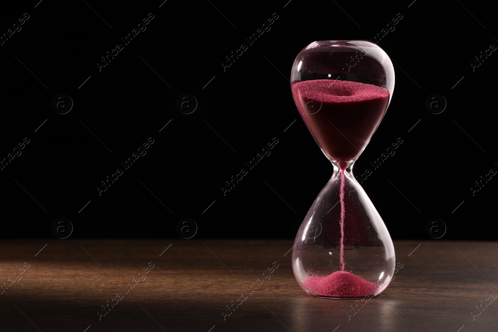 Photo of Hourglass with flowing sand on wooden table against black background. Space for text