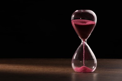 Photo of Hourglass with flowing sand on wooden table against black background. Space for text