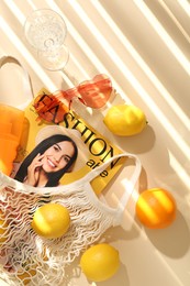 Photo of String bag with fresh fruits, fashion magazine and beach accessories on beige background, flat lay