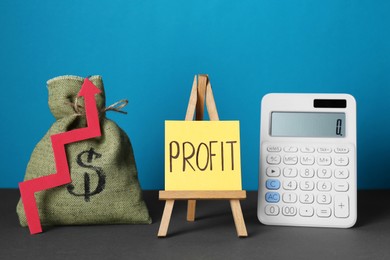 Photo of Economic profit. Money bag, arrow, calculator and easel with note on grey table against light blue background