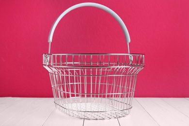 Photo of Empty metal shopping basket on white wooden table against crimson background