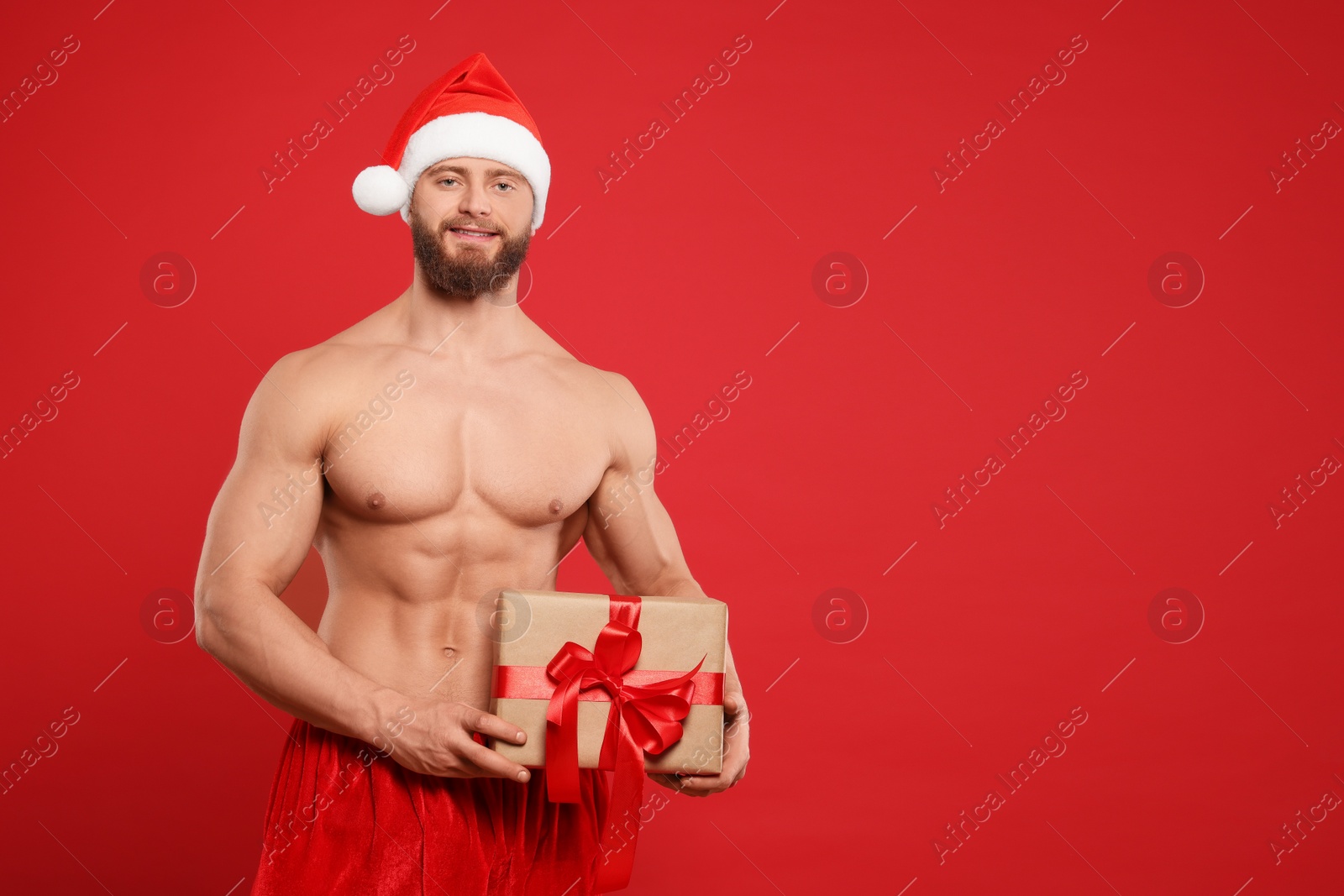 Photo of Attractive young man with muscular body in Santa hat holding Christmas gift box on red background, space for text