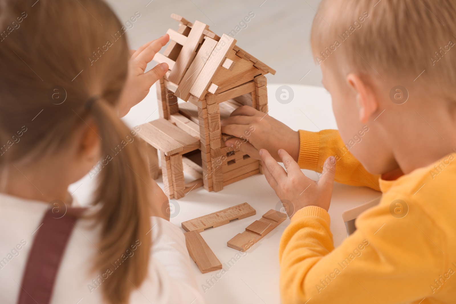 Photo of Little boy and girl playing with wooden house at white table indoors, closeup. Children's toys