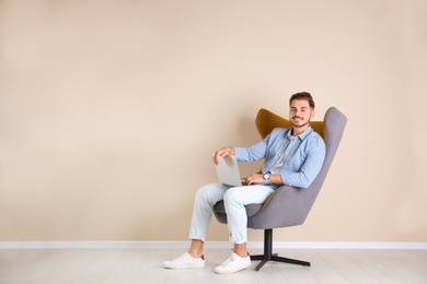 Handsome young man with laptop in armchair near color wall indoors. Space for text