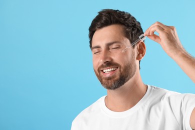 Smiling man applying cosmetic serum onto his face on light blue background. Space for text