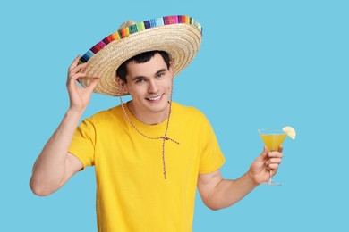 Young man in Mexican sombrero hat with cocktail on light blue background