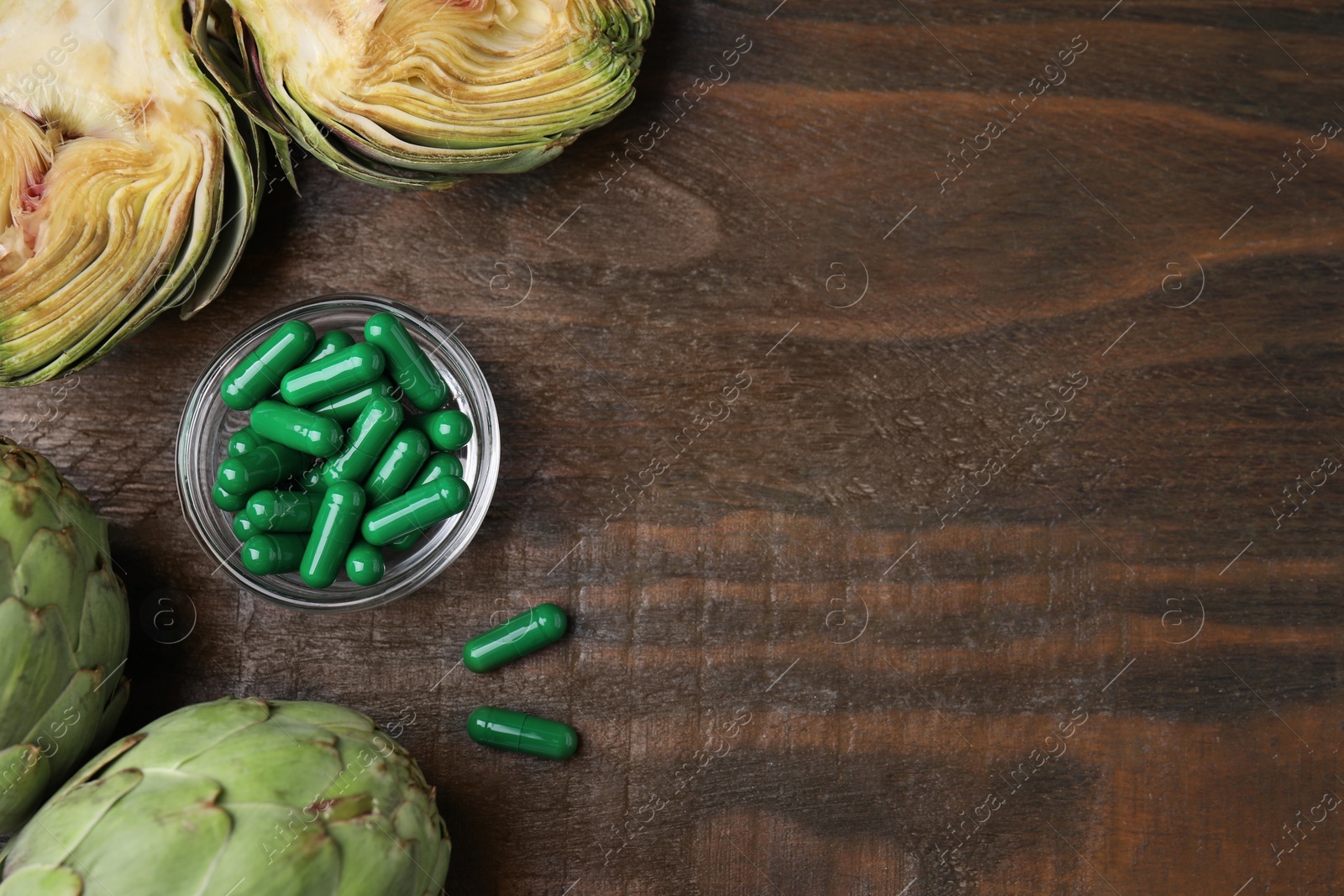 Photo of Bowl with pills and fresh artichokes on wooden table, flat lay. Space for text