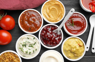 Photo of Different tasty sauces in bowls, tomatoes and spoons on black wooden table, flat lay