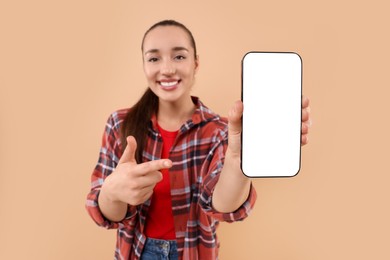 Photo of Young woman showing smartphone in hand and pointing at it on light brown background, selective focus. Mockup for design