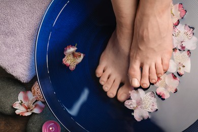 Photo of Woman soaking her feet in bowl with water and flowers on floor, top view. Spa treatment