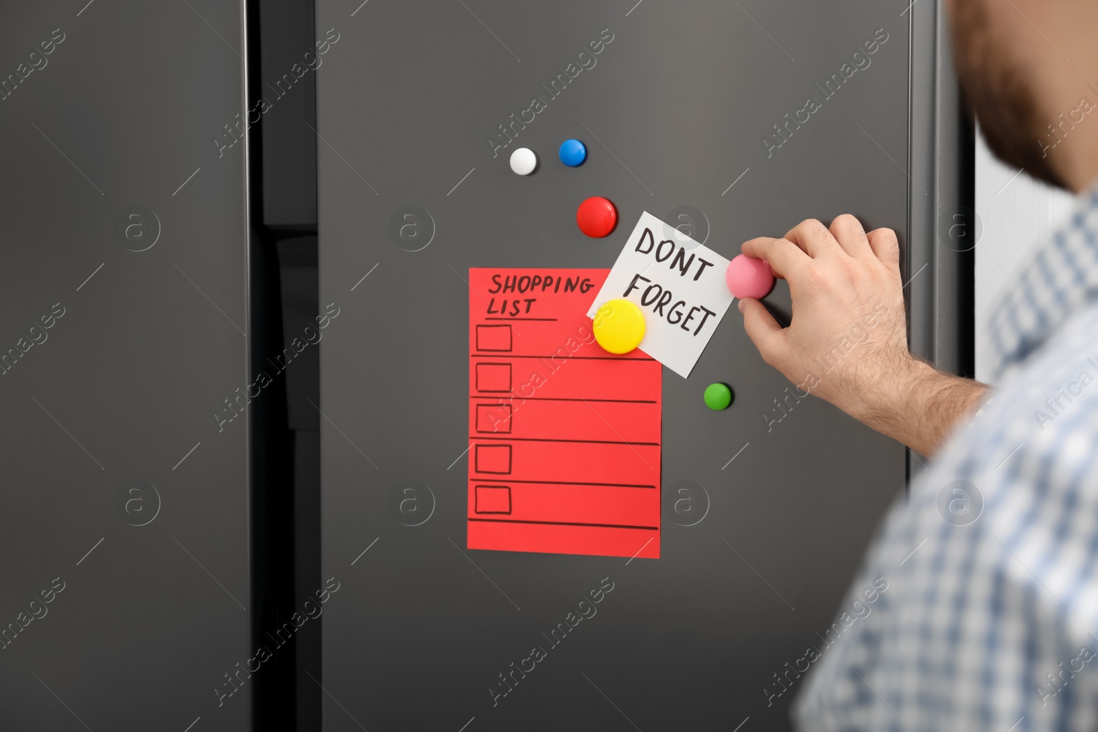 Photo of Man putting shopping list and reminder on refrigerator door, closeup