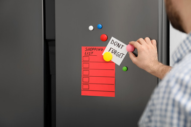 Photo of Man putting shopping list and reminder on refrigerator door, closeup