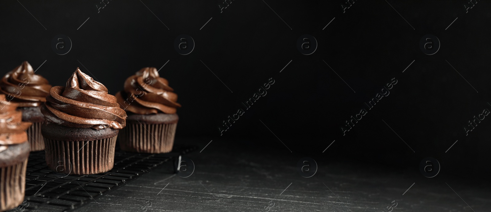 Photo of Cooling rack with delicious chocolate cupcakes on black table against dark background. Space for text