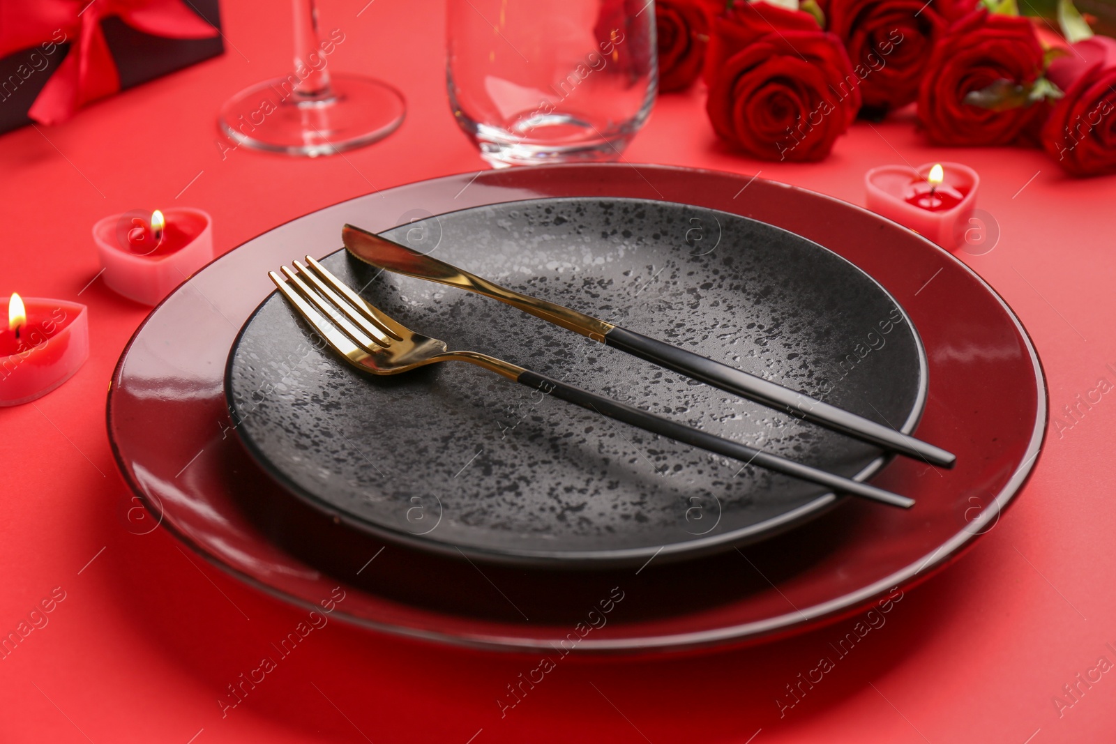 Photo of Place setting with heart shaped candles, gift box and bouquet of roses on red table, closeup. Romantic dinner