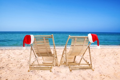 Image of Wooden deck chairs and Santa Claus hats on beach near sea. Christmas vacation
