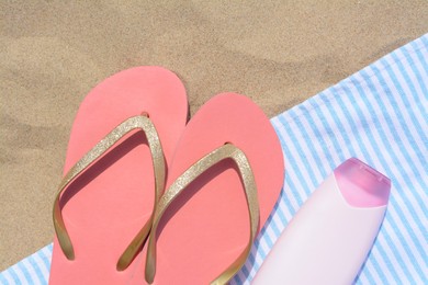 Photo of Striped towel with bottle of sunblock and flip flops on sandy beach, flat lay