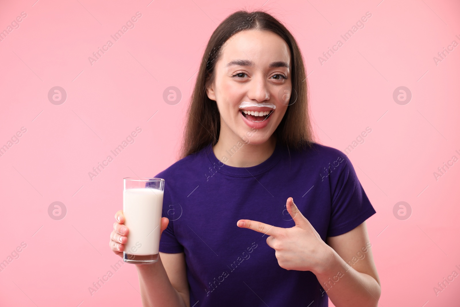 Photo of Happy woman with milk mustache pointing at glass of tasty dairy drink on pink background