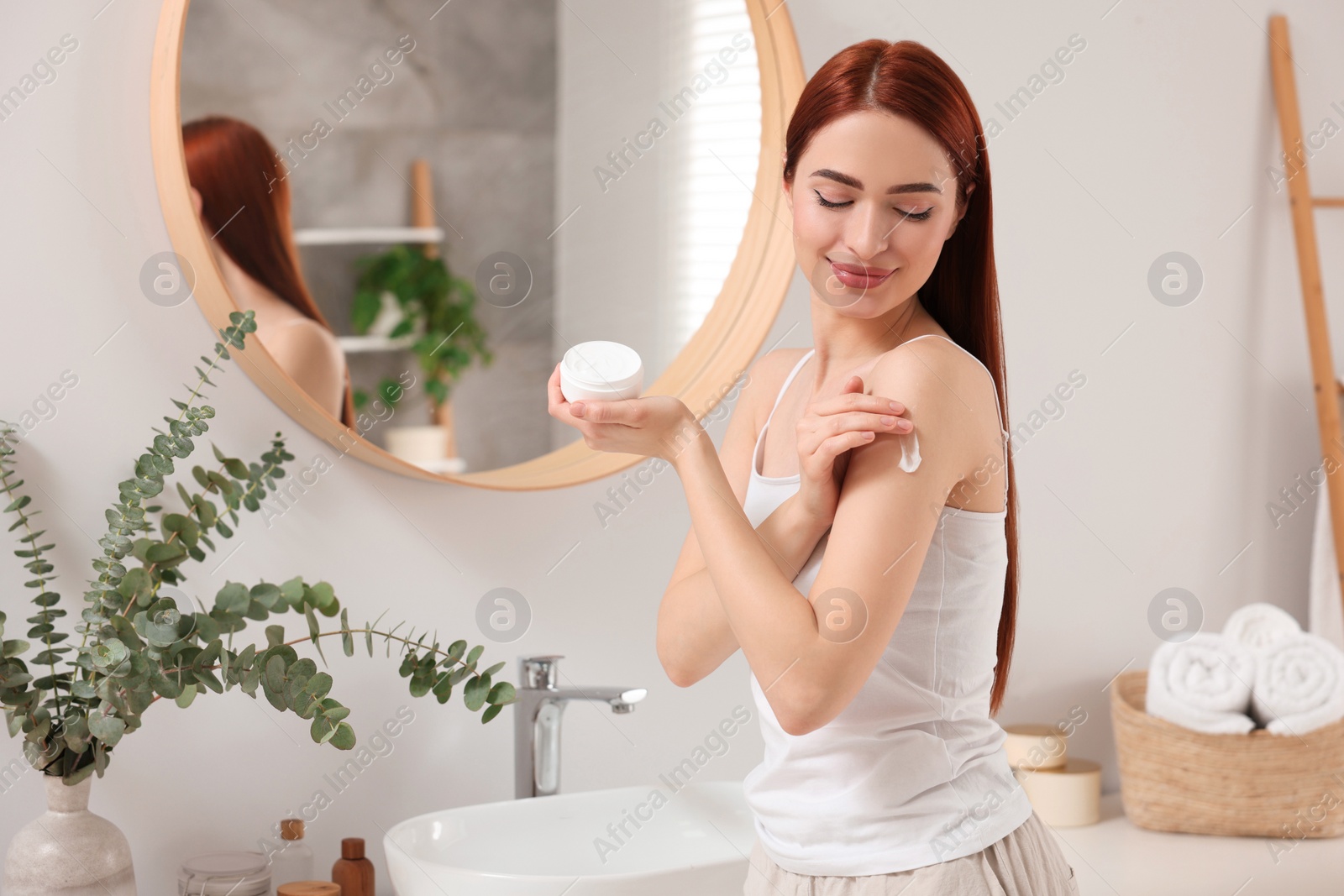 Photo of Beautiful young woman applying body cream onto shoulder in bathroom, space for text