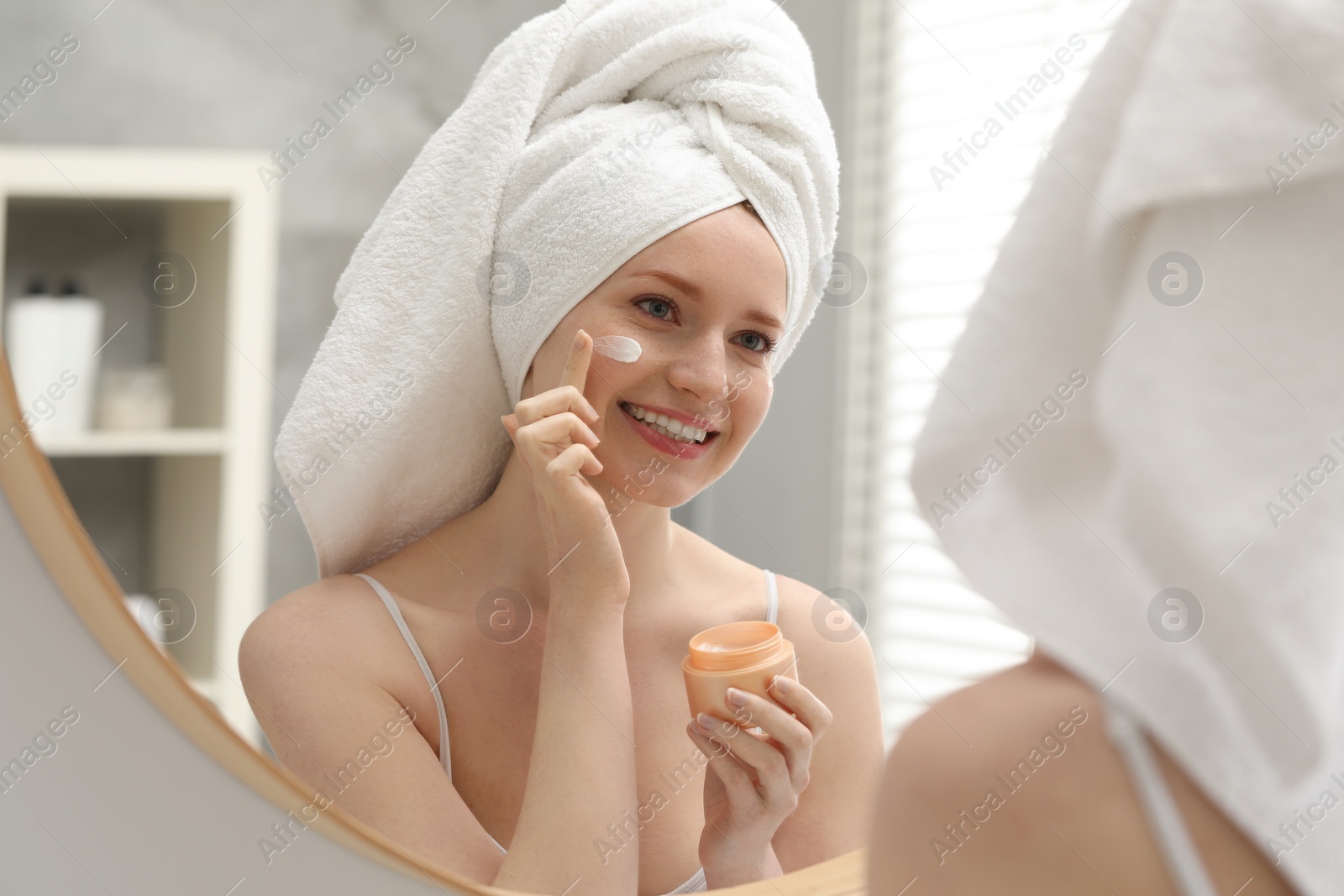 Photo of Smiling woman with freckles applying cream onto her face near mirror in bathroom