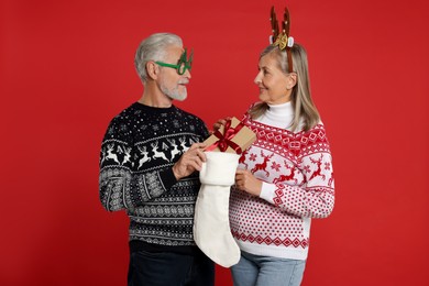 Photo of Senior couple in Christmas sweaters taking gift from stocking on red background