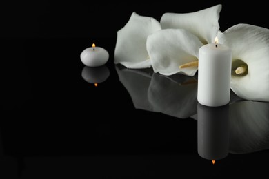 White calla lily flowers and burning candles on black mirror surface in darkness, space for text. Funeral symbols