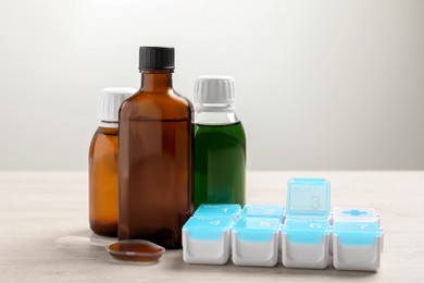 Bottles of syrup, dosing spoon and weekly pill organizer on white table against light grey background. Cold medicine