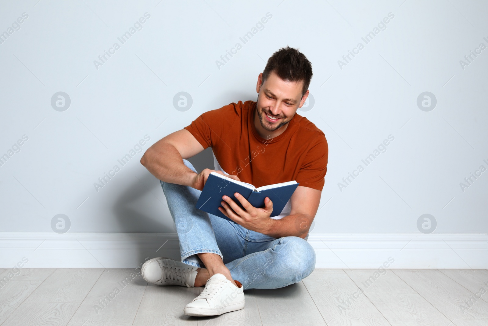 Photo of Handsome man reading book on wooden floor near light wall