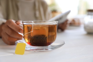 Woman holding glass cup with tea bag at white wooden table, closeup