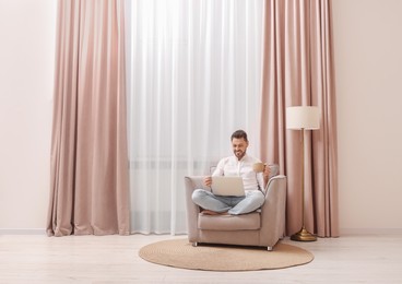 Photo of Smiling man drinking tea while working on laptop near window with beautiful curtains at home. Space for text