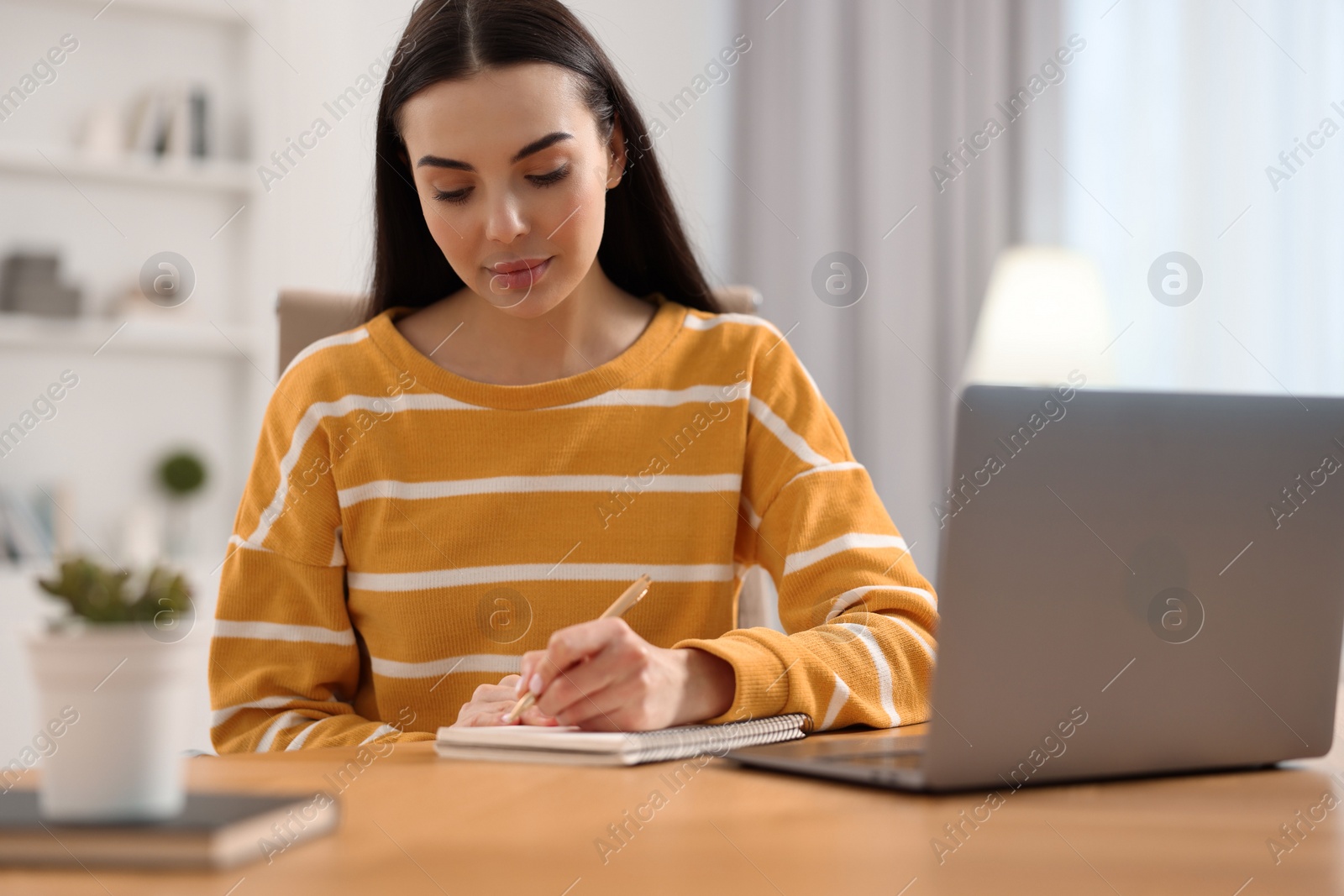 Photo of Young woman writing down notes during webinar at table in room