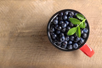 Tasty fresh bilberries with green leaves in mug on wooden table, top view. Space for text