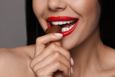 Young woman with red lips eating heart shaped chocolate candy on grey background, closeup